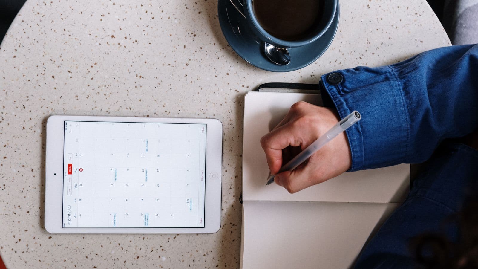 Calendário digital em um tablet ao lado de um caderno de anotações, caneta e xícara de café sobre a mesa